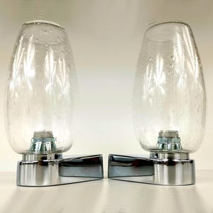 May include: A pair of wall-mounted light fixtures featuring clear, teardrop-shaped glass shades with a subtle bubble texture. Each shade is secured to a polished chrome base with a threaded metal fitting at the bottom of the glass.