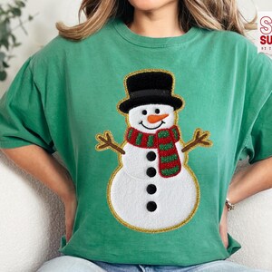 May include: A green t-shirt with a snowman design. The snowman has a black top hat, an orange nose, and a red and green striped scarf. The shirt has the text "DIGITAL DOWNLOAD" on the left side.