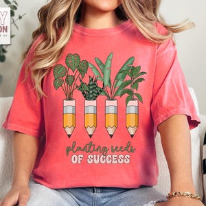 Peut inclure: T-shirt corail avec un motif de plantes dans des pots en forme de crayon. Le texte "planting seeds of success" est en dessous. Le t-shirt a des manches courtes et un col rond.