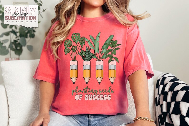 Peut inclure: T-shirt corail avec un motif de plantes dans des pots en forme de crayon. Le texte "planting seeds of success" est en dessous. Le t-shirt a des manches courtes et un col rond.