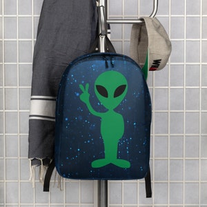 May include: A blue backpack with a green alien graphic on it. The alien is waving with one hand and has a peace sign with the other. The background is a dark blue with white stars.