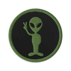 May include: Green alien with large eyes and a peace sign, embroidered on a black circle with a green outer ring.