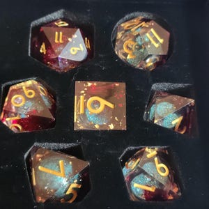May include: A set of seven polyhedral dice in a black box. The dice are a mix of red and clear resin with gold numbers and blue and gold glitter. The dice include a D20, D12, D10, D8, D6, D4, and a square D6.