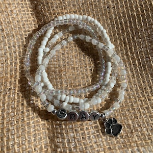 May include: A set of four white beaded bracelets with a silver charm that says "PRAY" and a paw print charm.