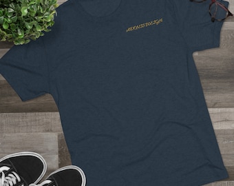 May include: A navy blue t-shirt with the embroidered text "ADONISTWEIGH" in gold. The shirt is displayed on a wooden surface, with a pair of black and white sneakers and a pair of eyeglasses.