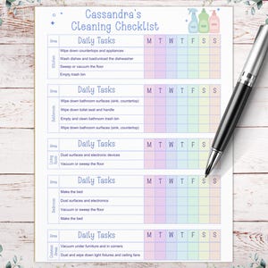 May include: A printable cleaning checklist titled "Cassandra's Cleaning Checklist" with daily tasks for the kitchen, bathroom, living room, bedroom, and common areas. A black and silver pen rests on the checklist. The checklist has a pastel rainbow color scheme.