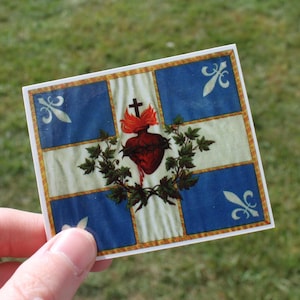 May include: A square sticker featuring a design with a central heart, cross, and flames, surrounded by a wreath. The background has blue squares with fleur-de-lis symbols and a cream-colored center.