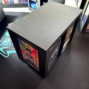 Puede incluir: Un expositor rectangular negro con superficie superior texturizada, dise&ntilde;ado para albergar y mostrar dos tarjetas coleccionables en fundas protectoras de pl&aacute;stico. La caja cuenta con dos ranuras verticales que enmarcan las tarjetas.