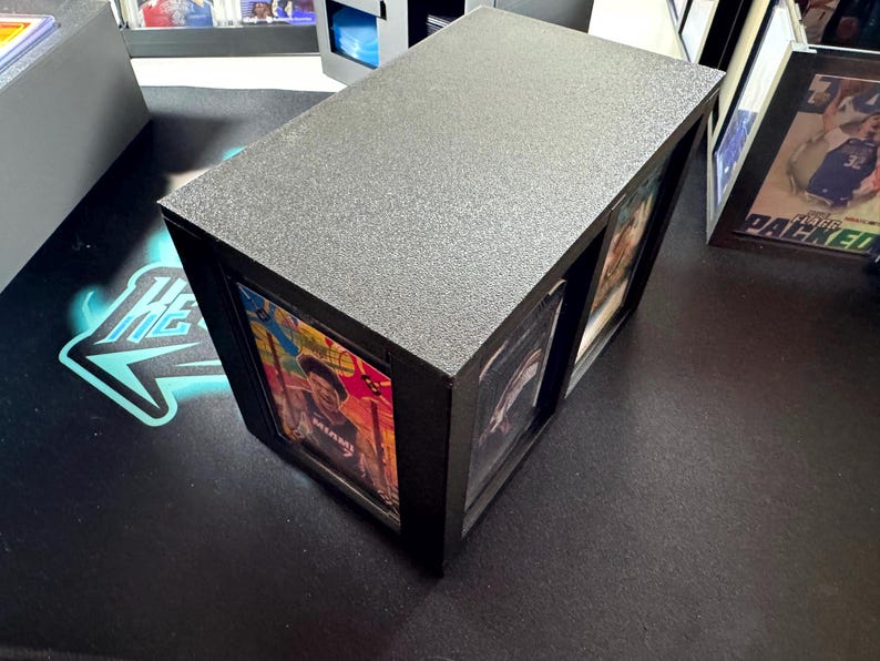 Puede incluir: Un expositor rectangular negro con superficie superior texturizada, dise&ntilde;ado para albergar y mostrar dos tarjetas coleccionables en fundas protectoras de pl&aacute;stico. La caja cuenta con dos ranuras verticales que enmarcan las tarjetas.