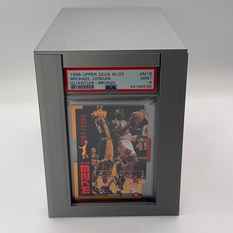 May include: A silver-colored display case holding a Michael Jordan basketball card. The card is from the 1998 Upper Deck MJ23 series, labeled "Quantum - Bronze." The card is graded as Mint 9. The case has the text "M723" and "0981/2300".