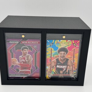 Puede incluir: Una vitrina rectangular negra que contiene dos tarjetas deportivas en soportes magn&eacute;ticos protectores. La tarjeta izquierda muestra a un jugador con camiseta roja y el texto New Recruits, la derecha a un jugador con camiseta de Miami.
