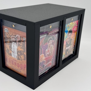 Puede incluir: Una vitrina rectangular negra con tres ranuras verticales que contienen tarjetas deportivas en fundas protectoras de pl&aacute;stico. Las tarjetas muestran jugadores de baloncesto, con texto visible que incluye Rookie Roundup y New Recruits detr&aacute;s de paneles transparentes.