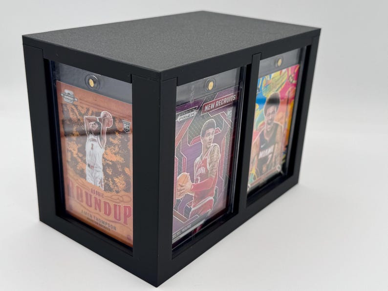 Puede incluir: Una vitrina rectangular negra con tres ranuras verticales que contienen tarjetas deportivas en fundas protectoras de pl&aacute;stico. Las tarjetas muestran jugadores de baloncesto, con texto visible que incluye Rookie Roundup y New Recruits detr&aacute;s de paneles transparentes.