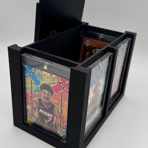 Puede incluir: Una caja de almacenamiento rectangular negra con tapa abierta, dise&ntilde;ada para guardar varias tarjetas coleccionables en fundas magn&eacute;ticas protectoras. El frente tiene una ranura que muestra una tarjeta de baloncesto colorida.