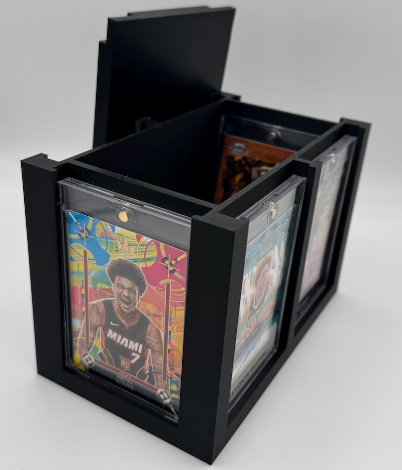 Puede incluir: Una caja de almacenamiento rectangular negra con tapa abierta, dise&ntilde;ada para guardar varias tarjetas coleccionables en fundas magn&eacute;ticas protectoras. El frente tiene una ranura que muestra una tarjeta de baloncesto colorida.