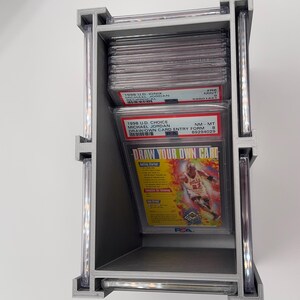 May include: A silver-colored, three-tiered display case holding graded sports cards. The cards feature Michael Jordan, with text including "1998 U.D. Choice" and "Draw Your Own Card." The case is designed to showcase and protect collectible cards.