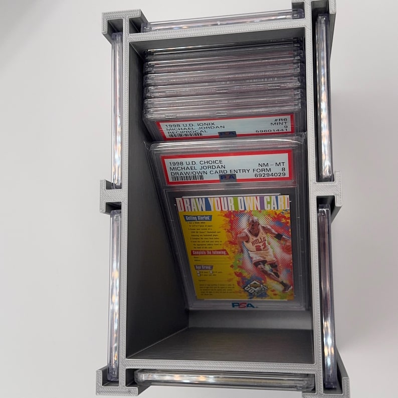 May include: A silver-colored, three-tiered display case holding graded sports cards. The cards feature Michael Jordan, with text including "1998 U.D. Choice" and "Draw Your Own Card." The case is designed to showcase and protect collectible cards.