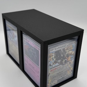 May include: A black, rectangular card display case with a textured finish. The case has three clear panels displaying collectible cards. The cards are visible through the clear panels. The case is designed to protect and showcase trading cards.