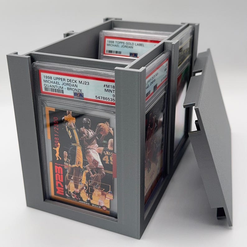 May include: A gray, rectangular card holder with a hinged lid, filled with graded sports cards. The cards feature Michael Jordan, with text including "1998 Upper Deck MJ23" and "1998 Topps Gold Label".