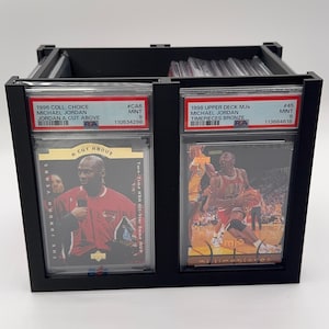 May include: Black display case holding two Michael Jordan trading cards. The cards are in protective cases and feature images of Michael Jordan. Text on the cards includes "1996 Coll. Choice" and "1998 Upper Deck MJx".