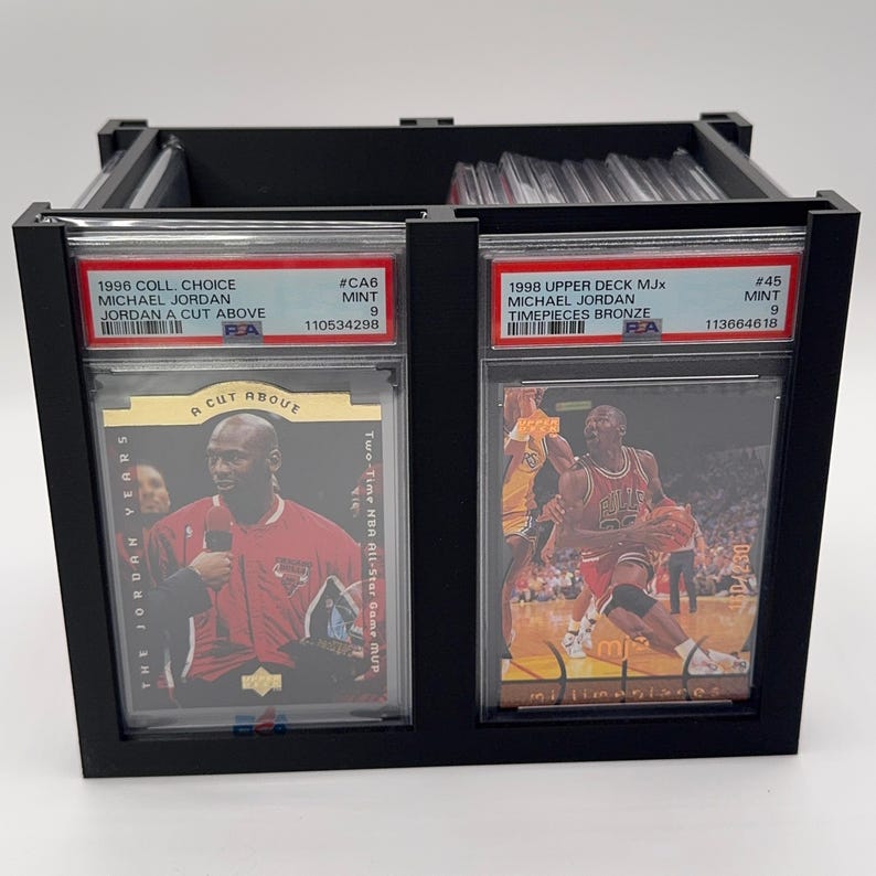 May include: Black display case holding two Michael Jordan trading cards. The cards are in protective cases and feature images of Michael Jordan. Text on the cards includes "1996 Coll. Choice" and "1998 Upper Deck MJx".