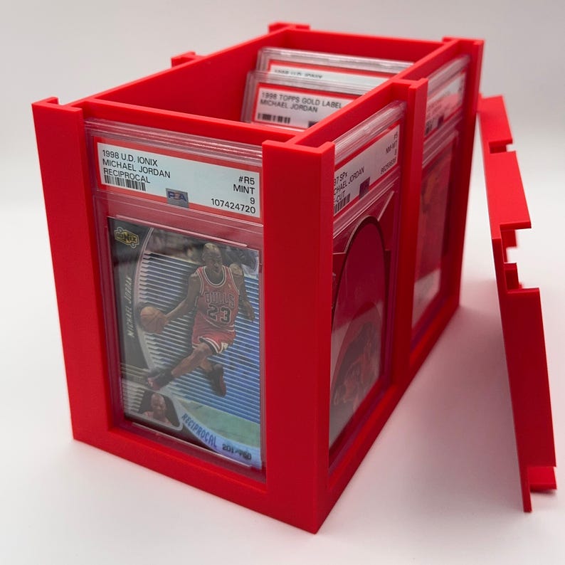 May include: A bright red card storage box with a removable lid. The box holds several graded sports cards, including a 1998 U.D. Ionix Michael Jordan card. The cards are in protective cases. The box is designed to keep cards organized.