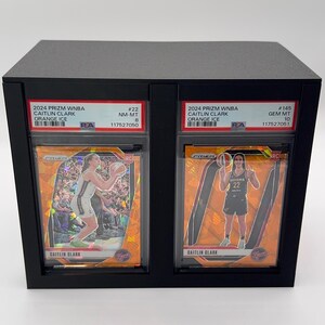 May include: A black display case holding two Caitlin Clark basketball trading cards. The cards are labeled "2024 Prizm WNBA Orange Ice" and feature action shots of the player. The cards are graded and encased.
