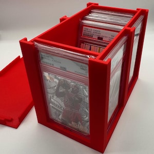 May include: A bright red trading card storage box, open to reveal graded cards. The box is rectangular with dividers and a removable lid. The cards feature Michael Jordan. Text on the cards includes "1998 UD CHOICE MICHAEL JORDAN" and "1995 METAL MICHAEL JORDAN".