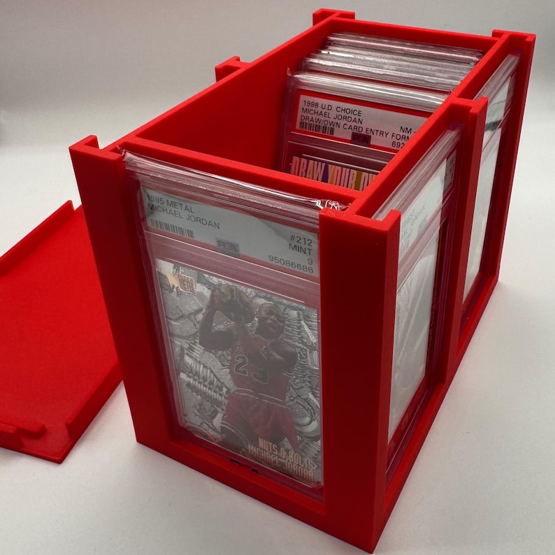 May include: A bright red trading card storage box, open to reveal graded cards. The box is rectangular with dividers and a removable lid. The cards feature Michael Jordan. Text on the cards includes "1998 UD CHOICE MICHAEL JORDAN" and "1995 METAL MICHAEL JORDAN".