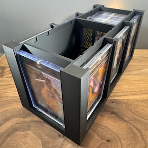 May include: A black 3D-printed storage organizer for trading cards, featuring multiple vertical slots designed to hold individual cards encased in clear plastic protective sleeves. The structure is rectangular with open compartments for display.