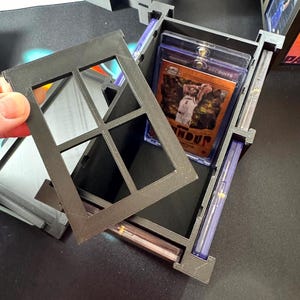 Puede incluir: Una caja de almacenamiento negra impresa en 3D para tarjetas coleccionables con una tapa extra&iacute;ble estilo ventana. La caja cuenta con ranuras internas dise&ntilde;adas para guardar varias tarjetas en fundas protectoras de pl&aacute;stico.