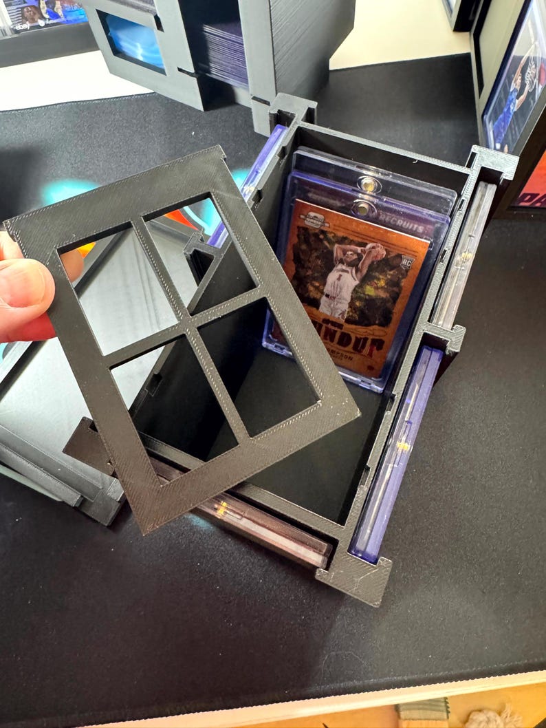 Puede incluir: Una caja de almacenamiento negra impresa en 3D para tarjetas coleccionables con una tapa extra&iacute;ble estilo ventana. La caja cuenta con ranuras internas dise&ntilde;adas para guardar varias tarjetas en fundas protectoras de pl&aacute;stico.