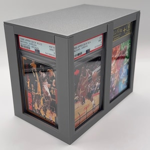 May include: A gray display case holding three graded basketball cards. The cards feature Michael Jordan and are labeled with details like "1998 Upper Deck MJ23" and "MINT 9". The case has a textured top and a sleek, modern design.