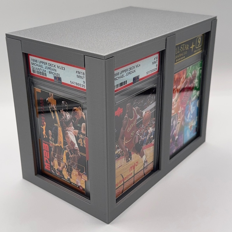 May include: A gray display case holding three graded basketball cards. The cards feature Michael Jordan and are labeled with details like "1998 Upper Deck MJ23" and "MINT 9". The case has a textured top and a sleek, modern design.