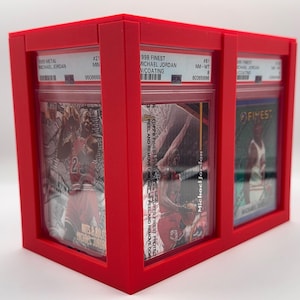 May include: A red display case holding three graded Michael Jordan trading cards. The cards are in protective cases, with visible labels and player images. The case is made of red material.
