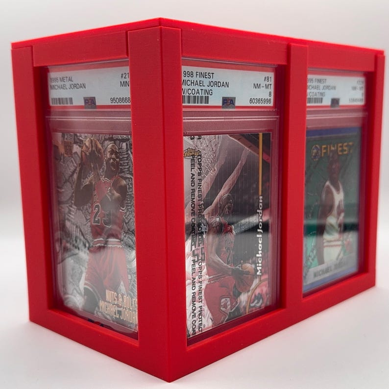 May include: A red display case holding three graded Michael Jordan trading cards. The cards are in protective cases, with visible labels and player images. The case is made of red material.