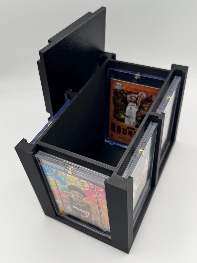 Puede incluir: Una caja de almacenamiento negra impresa en 3D para tarjetas coleccionables, con una ranura vertical para una tarjeta de exhibici&oacute;n y un compartimento interior abierto. La caja incluye una tapa con bisagras y fundas de pl&aacute;stico.