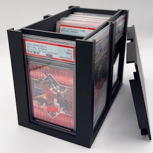 May include: Black display case holding graded sports cards. The case has a rectangular shape with a removable lid. The cards are visible through clear plastic sleeves, featuring Michael Jordan. The text on the cards reads "1999 Upper Deck Michael Jordan Athlete of the Century".