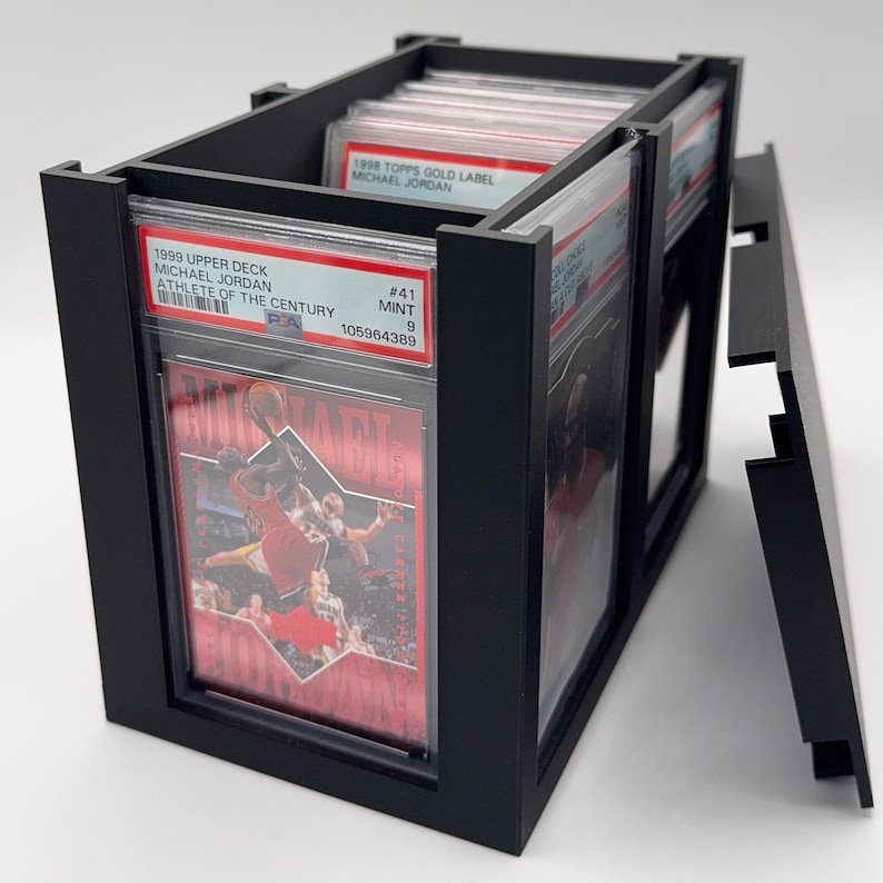 May include: Black display case holding graded sports cards. The case has a rectangular shape with a removable lid. The cards are visible through clear plastic sleeves, featuring Michael Jordan. The text on the cards reads "1999 Upper Deck Michael Jordan Athlete of the Century".