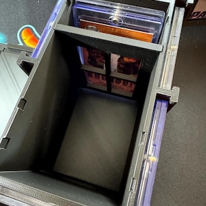Puede incluir: Una caja de almacenamiento negra impresa en 3D para tarjetas coleccionables, con un marco rectangular y divisores internos. La caja contiene varios estuches de pl&aacute;stico transparente, con uno visible en el compartimento principal y otros en los laterales.