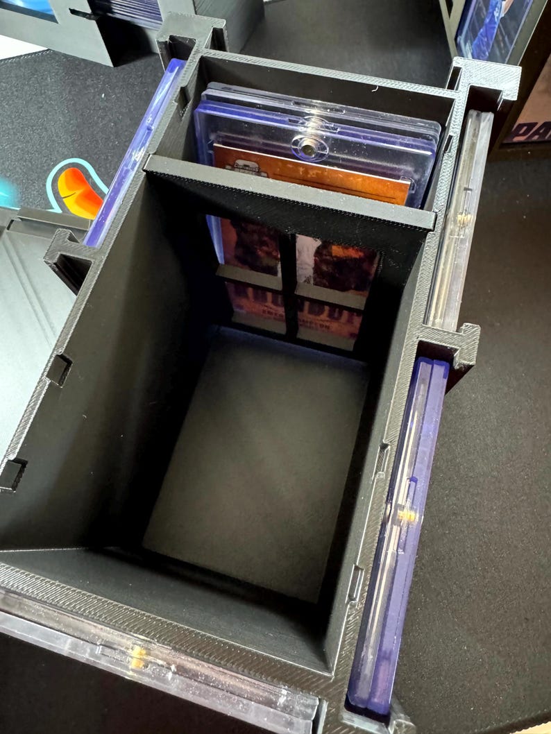 Puede incluir: Una caja de almacenamiento negra impresa en 3D para tarjetas coleccionables, con un marco rectangular y divisores internos. La caja contiene varios estuches de pl&aacute;stico transparente, con uno visible en el compartimento principal y otros en los laterales.