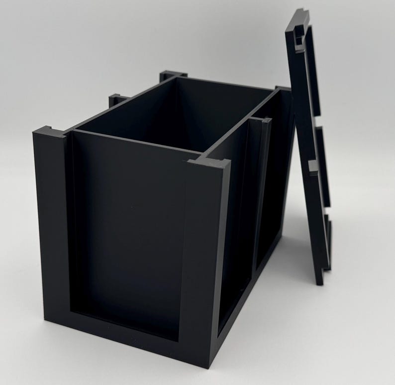 Puede incluir: Un contenedor de almacenamiento rectangular de color negro mate con la parte superior abierta y una tapa a juego separada colocada verticalmente al lado. El contenedor presenta un dise&ntilde;o estructurado con paredes gruesas y bordes con muescas.