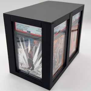 May include: Black display case holding graded sports cards. The case features a rectangular design with a top and front opening. The cards are visible through clear protective sleeves. Text on the cards includes "1998 TOPPS CHROME MICHAEL JORDAN CHAMPION SPIRIT".