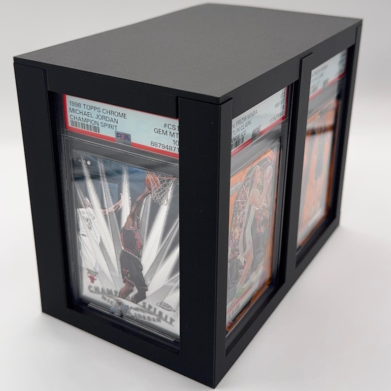 May include: Black display case holding graded sports cards. The case features a rectangular design with a top and front opening. The cards are visible through clear protective sleeves. Text on the cards includes "1998 TOPPS CHROME MICHAEL JORDAN CHAMPION SPIRIT".