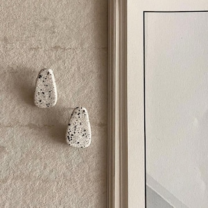 May include: Two stud earrings with a teardrop shape, featuring a speckled pattern of black dots on a cream-colored base. The earrings have a smooth, matte finish and are positioned next to the edge of a book with white pages and a black border.