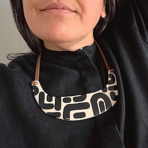 May include: A crescent-shaped statement necklace featuring a cream-colored base with a bold, black abstract pattern. The piece is suspended from a thin, brown leather cord that attaches to the sides of the curved, smooth-surfaced pendant.