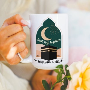 May include: White ceramic mug with a green and gold design featuring a crescent moon, a mosque, and the text "First Hajj Together Est. 1445 Maryam & Ali".