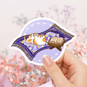 May include: A sticker featuring two cartoon cats resting on a purple prayer rug with yellow floral accents and a fringed edge. An open book on a wooden stand is next to the cats. The sticker is in a circular shape.