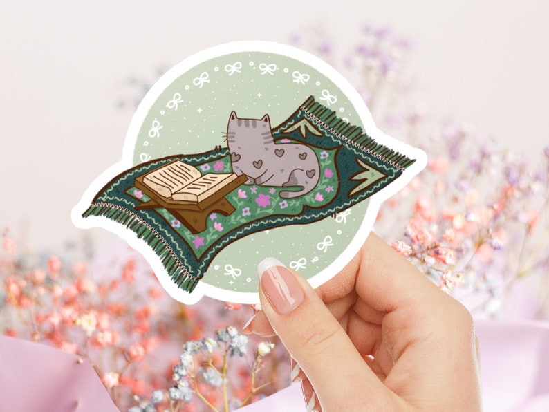 May include: A green prayer rug with a gray cat sitting on it. The rug has a floral pattern and a book is on a stand in front of the cat. The rug is on a light green background with white bows.