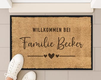 May include: A rectangular doormat with the text "WILLKOMMEN BEI Familie Becker" in brown lettering, with two hearts and a line underneath. The mat is brown with a black border. White sneakers are visible.